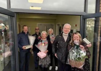 Six guests gathered in the centre doorway holding bouquets, gift boxes and a hamper.