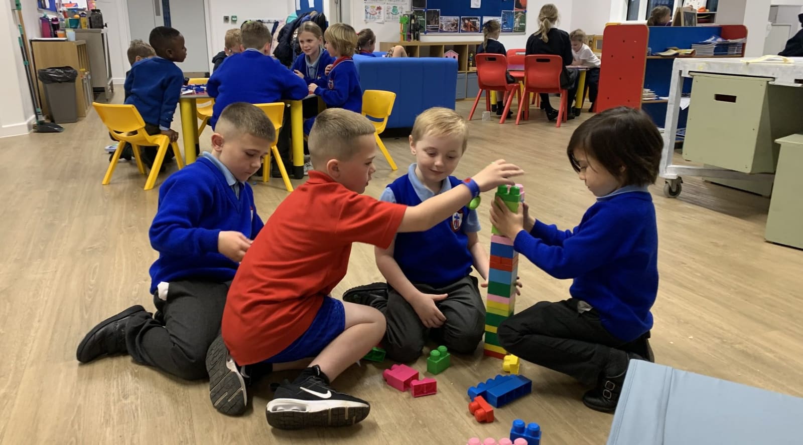Four primary school children in uniform kneeling on the floor, building a tall tower of coloured bricks together.