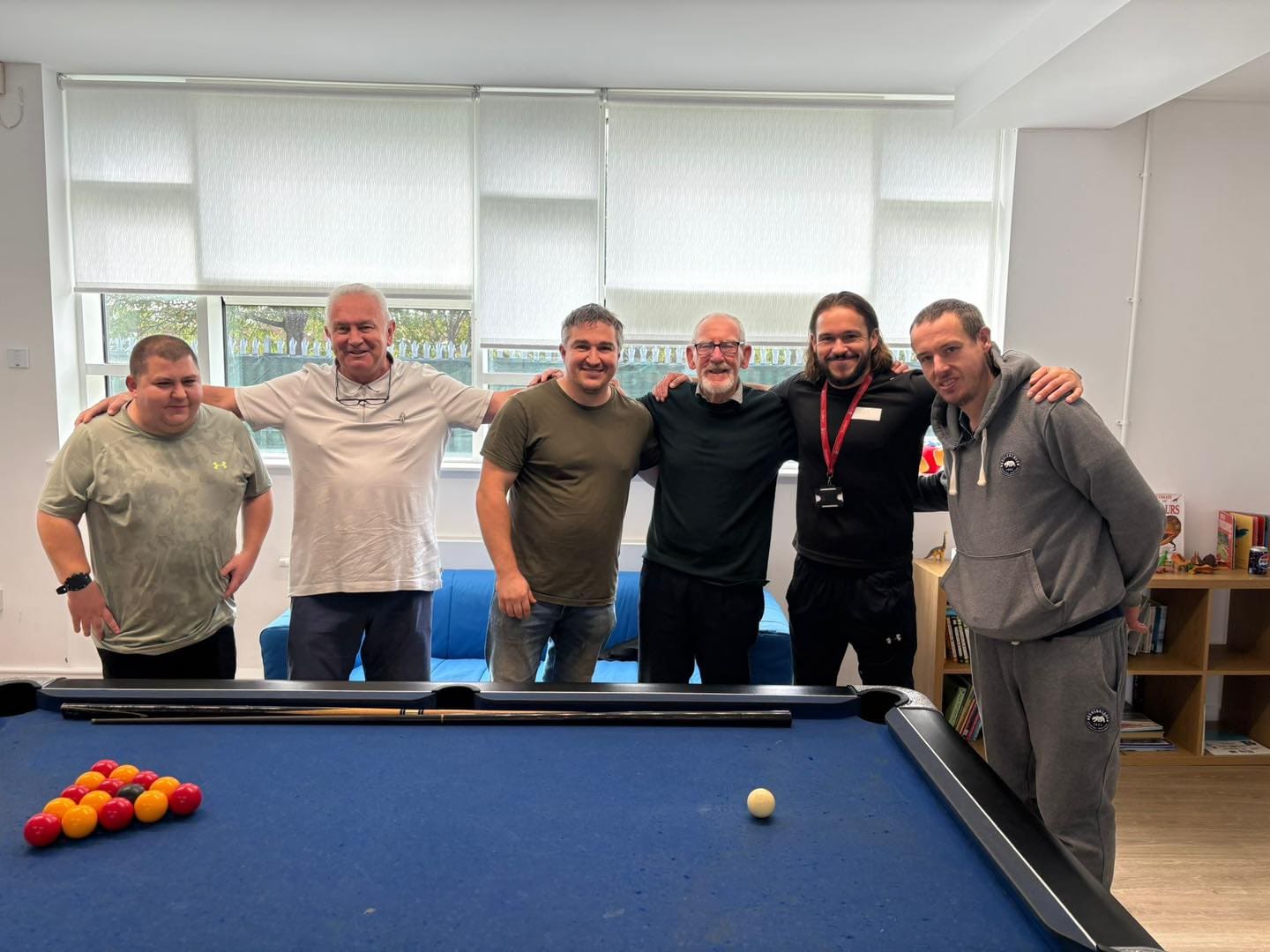 Six men with their arms around each other beside the pool table in the community centre lounge.