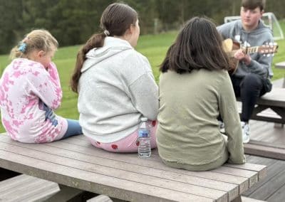 Three children sitting on a picnic bench while a youth worker plays guitar